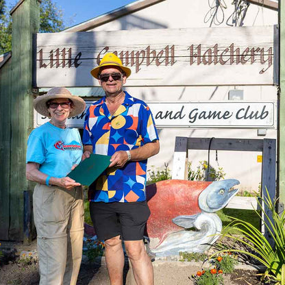 Two people standing outdoors in front of a wooden sign that reads "Little Campbell Hatchery" and "and Game Club," with a colorful fish sculpture below. The person on the right wears a bright geometric print shirt, yellow hat, and sunglasses, while the person on the left wears a light blue shirt and khaki pants.